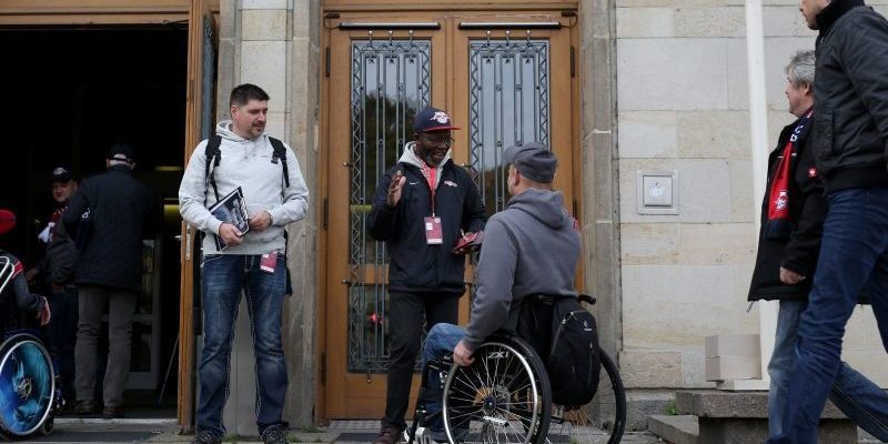 Mitarbeiter von RB Leipzig begrüßen einen Fan im Rollstuhl. Sie sind vor dem Eingang zu einem Raum mit der Aufschrift: RB Leipzig Club Scouts