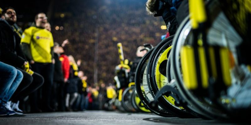 Borussia Dortmund Tribüne. Schwarz-gelbe Räder von Rollstühlen, dahinter Fans mit und ohne Trikots.