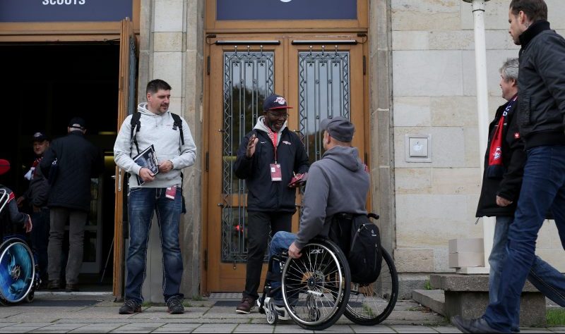 RB Leipzig Rollstuhlfahrer im Gespräch mit dunkelhäutigem Mitarbeiter der Behindertenbetreuung vor dem Eingang eines Fanraumes.