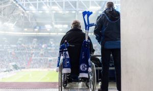 Rollstuhlfahrer mit Schalkeschals am den Griffen des Rollstuhls auf der Tribüne mit Blick auf das Spielfeld. Daneben ein stehender Fan.