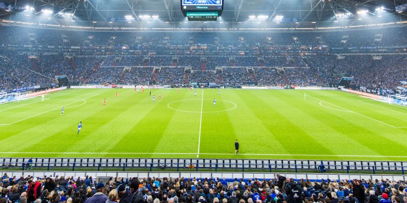 Blick auf das Spielfeld des Stadions von Schalke 04. Man sieht auch einen Teil der Zuschauer auf den Tribünen. Das Flutlicht ist an.