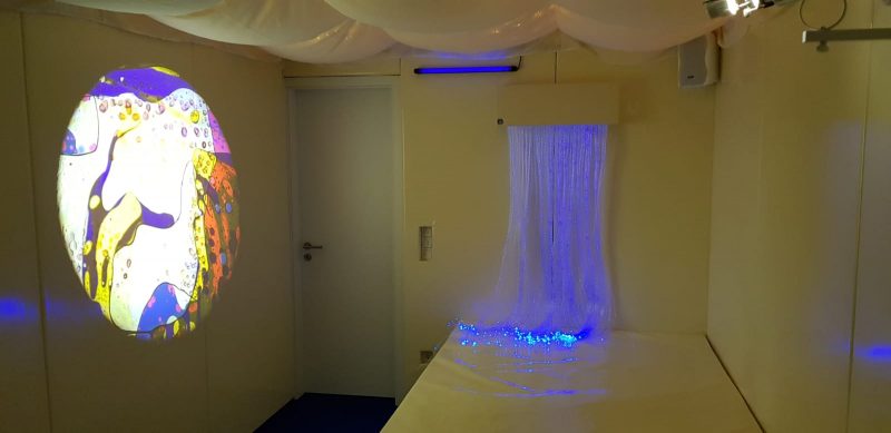 Snoezelraum. beruhigender Fasernebel leuchtet blau. Davor gibt es in Wasserbett zum Ausruhen, ein Bild in beruhigenden Farben wird an einer Wand angestrahlt.