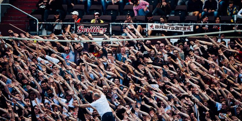Fans des FC St. Pauli bei einem Auswärtsspiel auf der Tribüne. Sie haben die Arme in die Höhe gestreckt und lehnen sich dabei zur Seite. Dahinter sitzen weitere Fans.