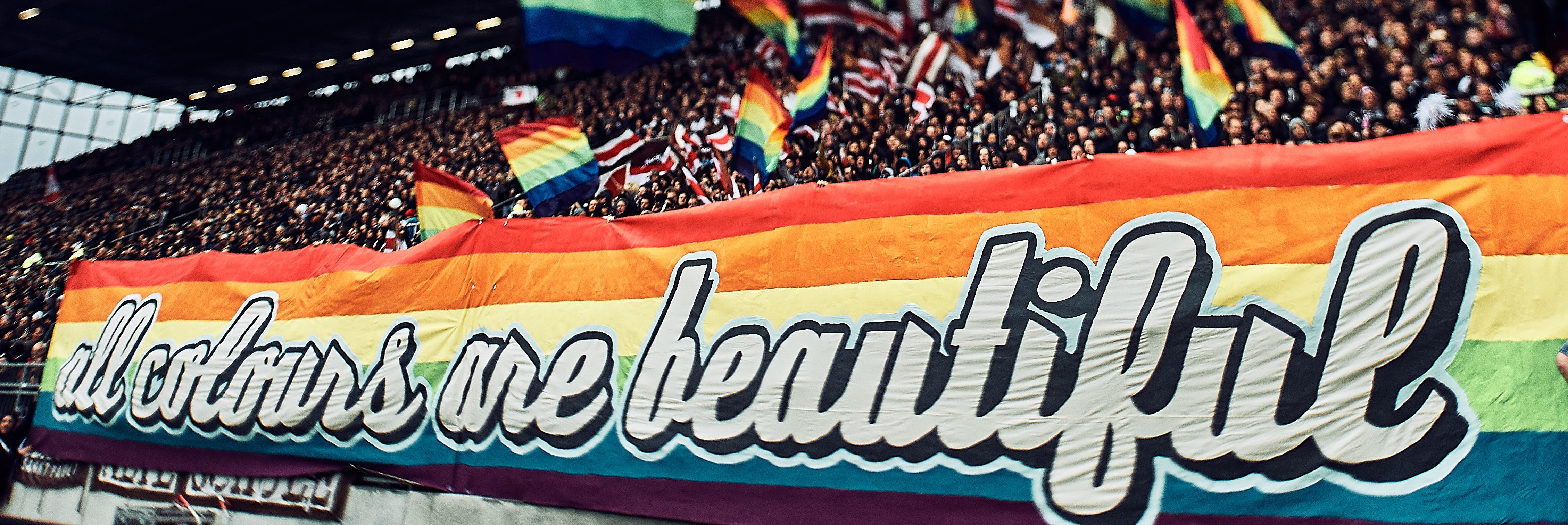Choreographie St. Pauli. Unten Banner in Regenbogenfarben mit Aufdruck "all colours are beautiful". Darüber schwingen Fans bunte Fahnen.