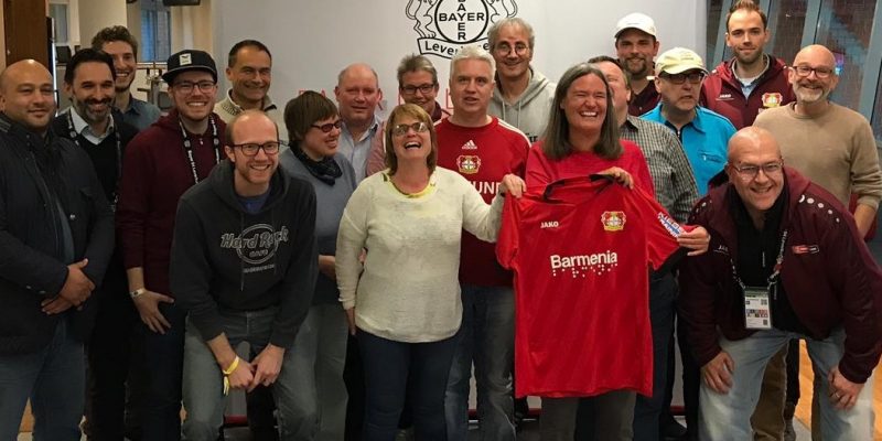 Team der Behindertenfanbetreuung und hörbehinderte Fans stehen vor einem Banner mit dem Logo von Bayer 04 Leverkusen, lachen in die Kamera. Eine Frau hält das Bayer Leverkusen Trikot mit der Brailleschrift auf dem Ärmel hoch.