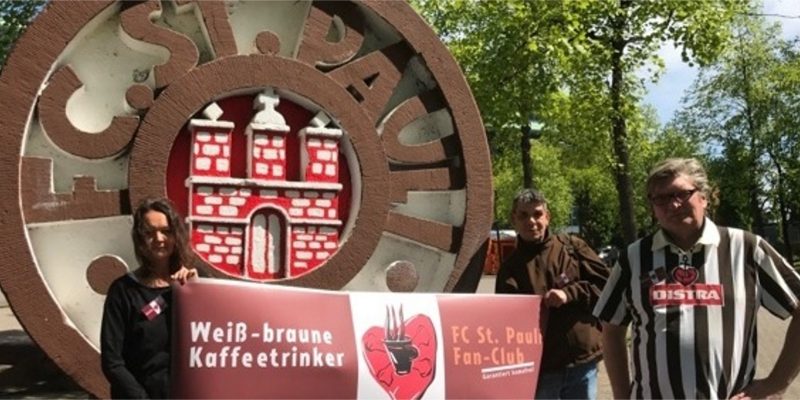 Drei Personen halten ein Banner der weiß braunen Kaffeetrinker*innen hoch. Sie stehen vor eine großen St. Pauli Logo