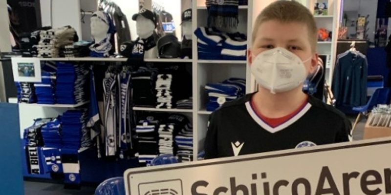 Der junge Arminia-Fan Paul mit einem "SchücoArena"-Schild und Mund-Nasen-Schutz während der "stillen Stunde" im Fanshop.
