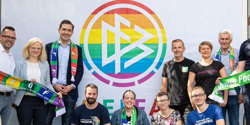 DFB-Logo in Regenbogenfarben auf einer Plakatwand. Davor versammelten sich Fans, die sich gegen Homophobie im Fußball engagieren.