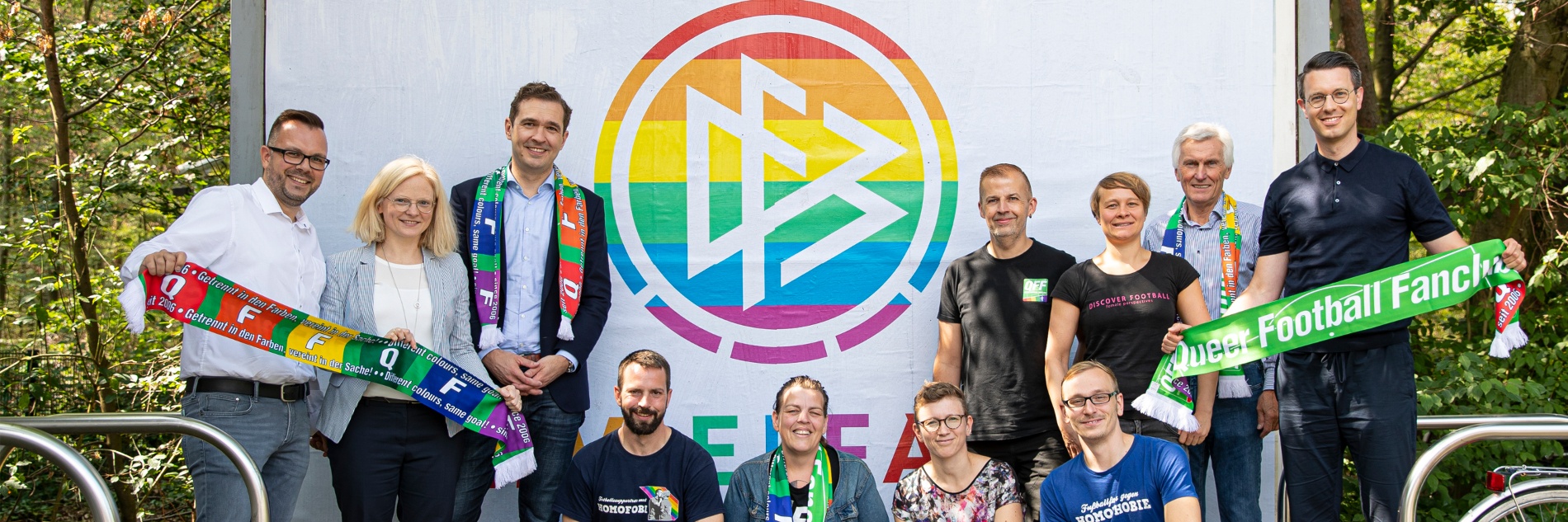 DFB-Logo in Regenbogenfarben auf einer Plakatwand. Davor versammelten sich Fans, die sich gegen Homophobie im Fußball engagieren.