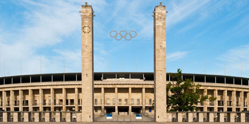 Der Haupteingang (Osttor) vom Olympiastadion Berlin vor blauem Himmel von außen fotografiert. Zwischen zwei Türmen sind die fünf olympischen Ringe sowie die Stadionränge von hinten zu sehen.