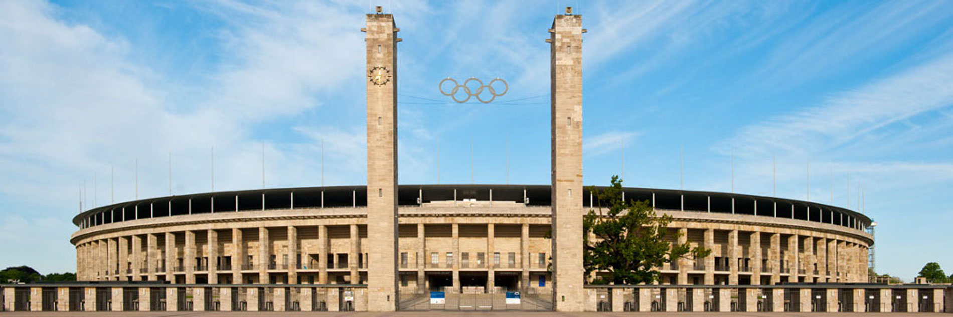 Der Haupteingang (Osttor) vom Olympiastadion Berlin vor blauem Himmel von außen fotografiert. Zwischen zwei Türmen sind die fünf olympischen Ringe sowie die Stadionränge von hinten zu sehen.