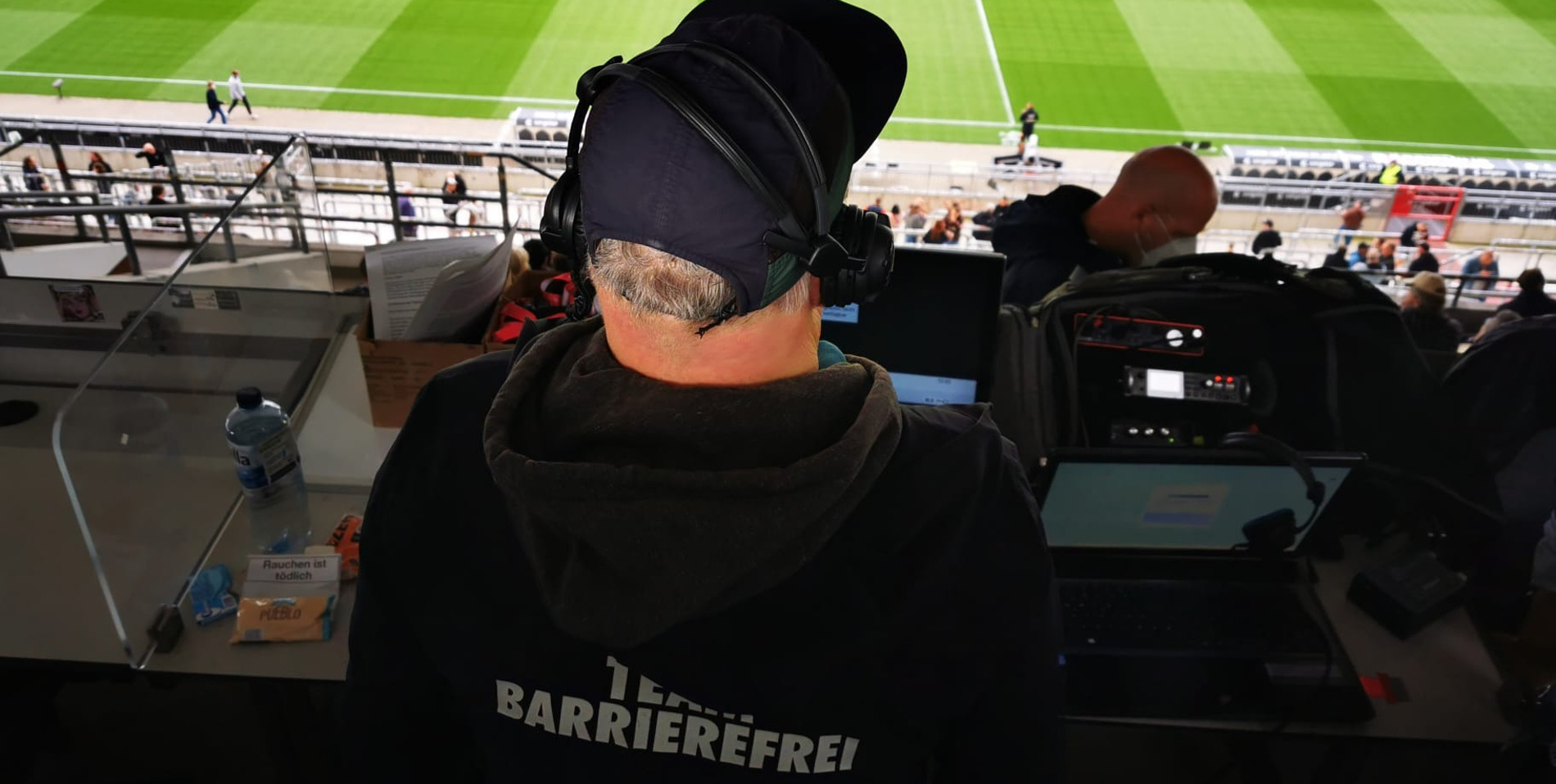 Person von hinten fotografiert, die einen Pullover mit Team Barrierefrei-Aufschrift und Kopfhörer trägt. Die Person guckt dabei auf das Spielfeld.