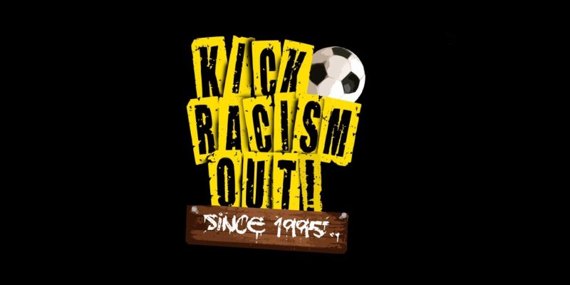 Gelbe-schwarzer "Kick Racism Out"-Schriftzug mit einem braun-weißen "Since 1995"-Schild in Holzoptik sowie einem schwarz-weißen Fußball garniert.