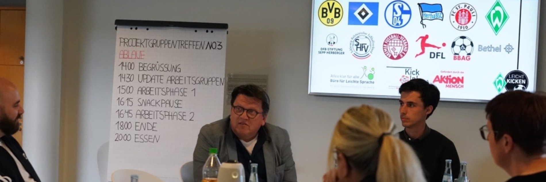 Die Teilnehmenden beim Projektgruppentreffen. Im Hintergrund das Programm des Treffens auf einem Flipchart. Auf einem TV werden die Wappen der beteiligten Clubs und Initiativen gezeigt.