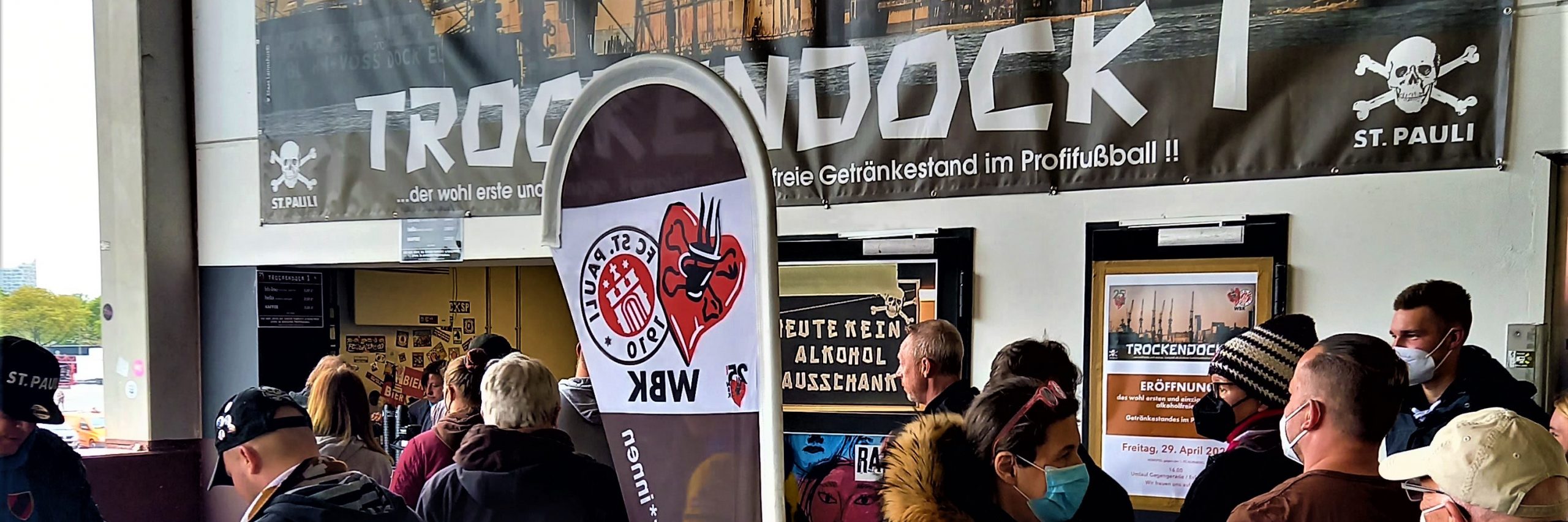 FC St. Pauli-Fans stehen vorm Trockendock I an. Davor eine Beachflag der Weiß-braune Kaffeetrinker*innen.