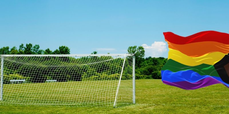 Die Regenbogenflagge ist die Flagge der „LGBTIAQ+“-Gemeinschaft vorm blauen Himmel. Daneben ein Fußballfeld.