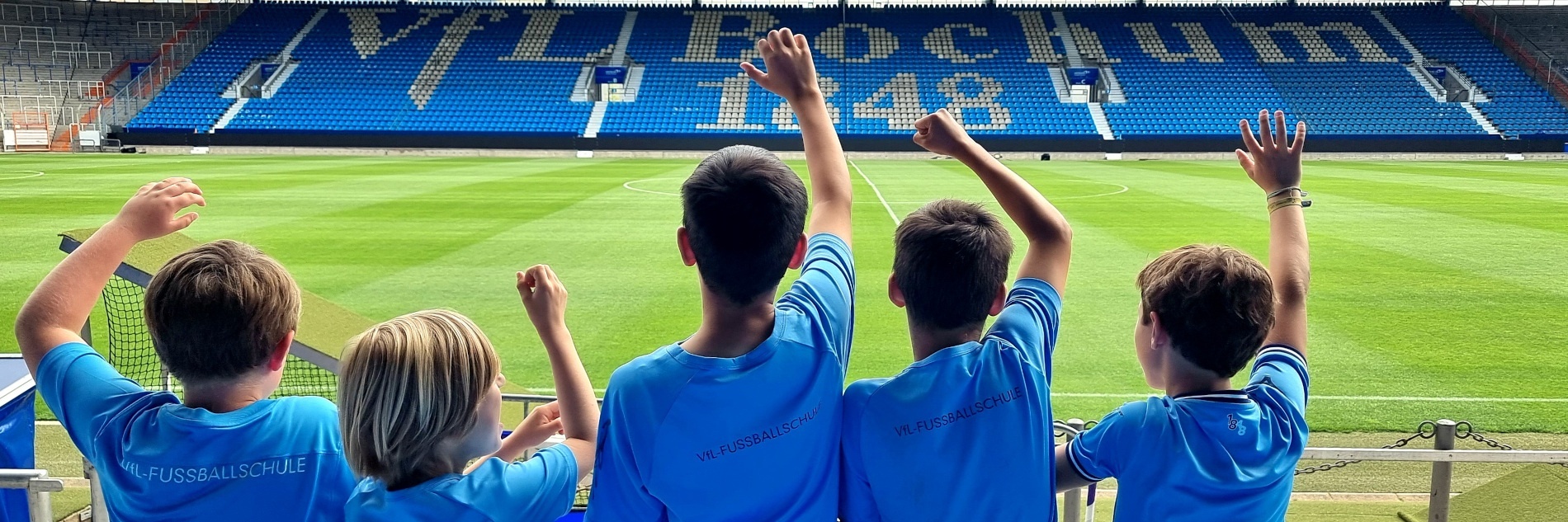 Eine Gruppe von Kindern mit hochgerissenen Armen von hinten fotografiert. Die Kinder schauen auf das Spielfeld und die leere Tribüne mit VfL Bochum 1848-Schriftzug im Hintergrund.