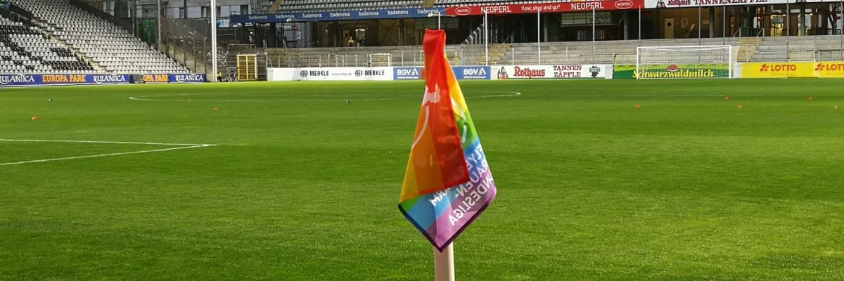 Eckfahne in Regenbogen-Farben mit Rasen und den Rängen des Dreisamstadions im Hintergrund. 