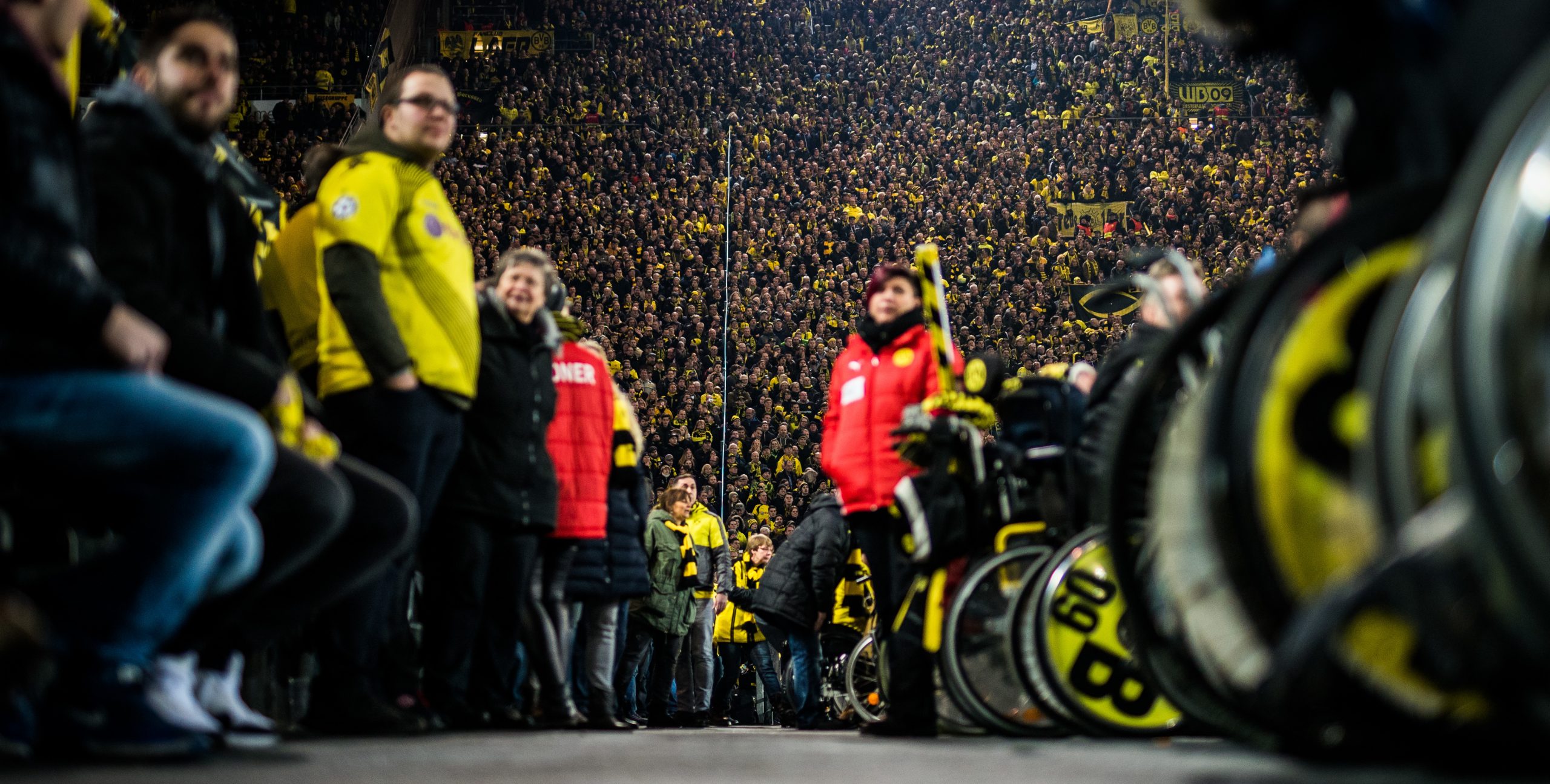 Stehende BVB-Fans links im Bild, in der Mitte ein Gang im Stadion und Ordner*innen, rechts BVB-Fans im Rollstuhl.