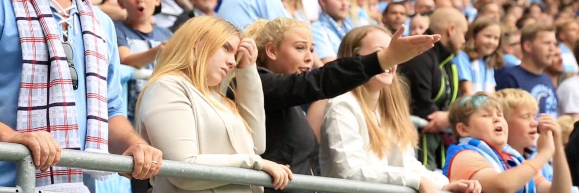 Drei Frauen stehen in einem Fußballstadion. Eine der Frauen streckt dabei ihre Hand in Richtung Spielfeld aus.