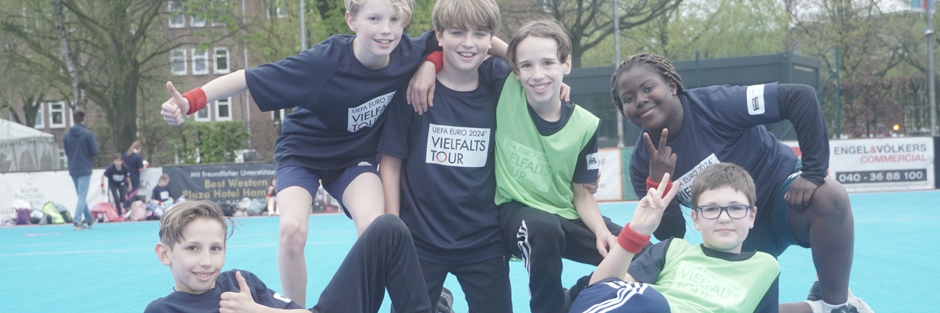 Sechs Kinder versammeln sich auf einem Sportplatz für ein Gruppenfoto und lächeln in die Kamera. Die Kinder tragen Trikots mit "UEFA EURO 20224 Vielfaltstour"-Aufschrift.
