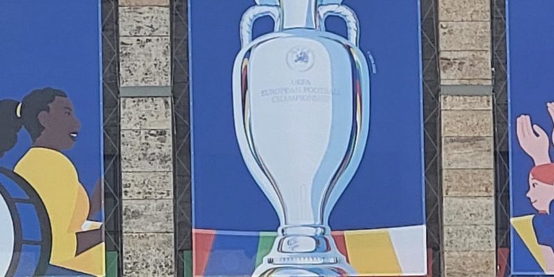 Buntes Branding der UEFA EURO 2024 mit dem Pokal der Europameisterschaft in der Mitte. Links davon sind zwei Fans mit einer Trommel, rechts sind Fans am Klatschen und mit Fahne.