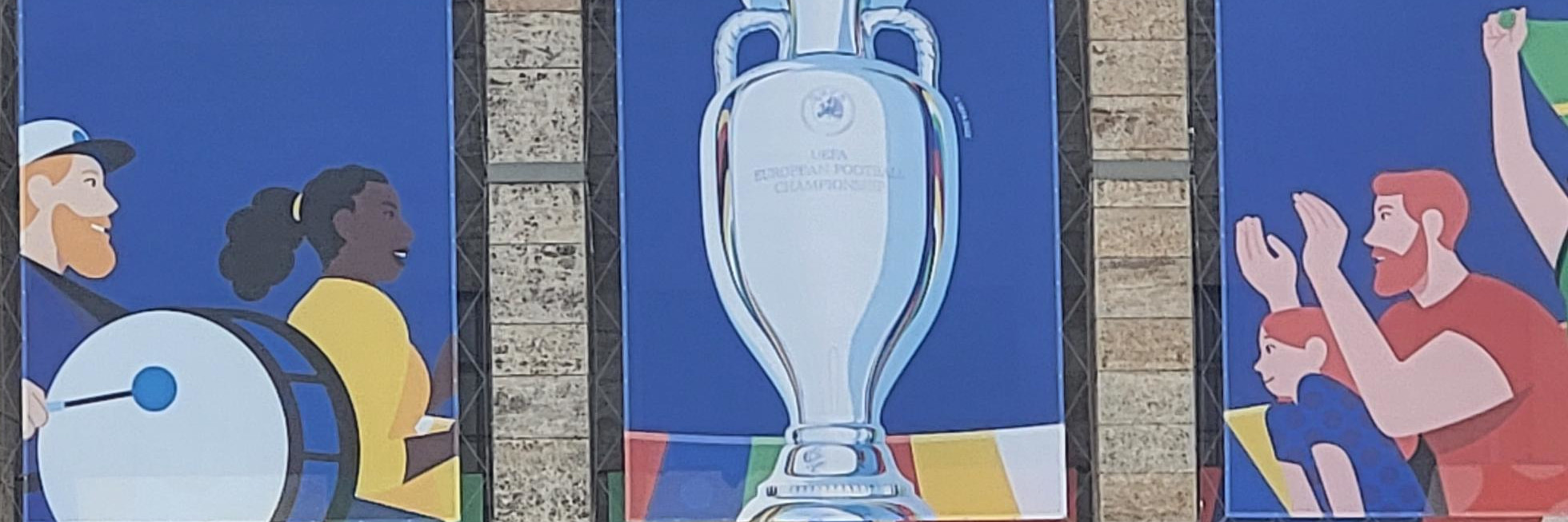 Buntes Branding der UEFA EURO 2024 mit dem Pokal der Europameisterschaft in der Mitte. Links davon sind zwei Fans mit einer Trommel, rechts sind Fans am Klatschen und mit Fahne.