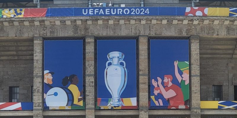 Blick auf die Außenfassade eines Stadions mit buntem Branding der EURO2024 inklusive einem Pokal sowie einem "UEFA EURO 2024"-Schriftzug.