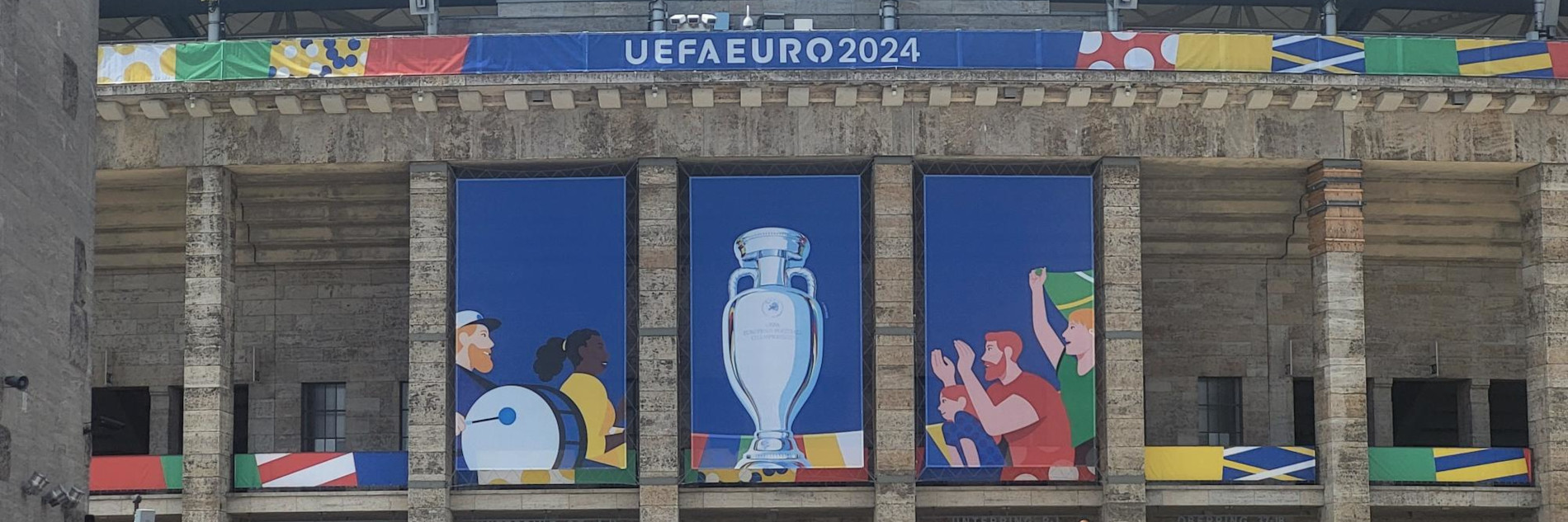 Blick auf die Außenfassade eines Stadions mit buntem Branding der EURO2024 inklusive einem Pokal sowie einem "UEFA EURO 2024"-Schriftzug.