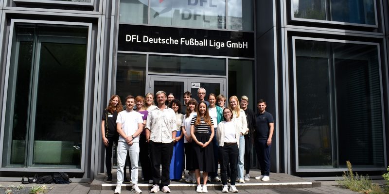 Eine Gruppe von 17 Menschen steht vor der DFL-Zentrale in Frankfurt. Über der Gruppe von Menschen stehen die Worte: “DFL Deutsche Fußball Liga GmbH” mit dem DFL-Logo.