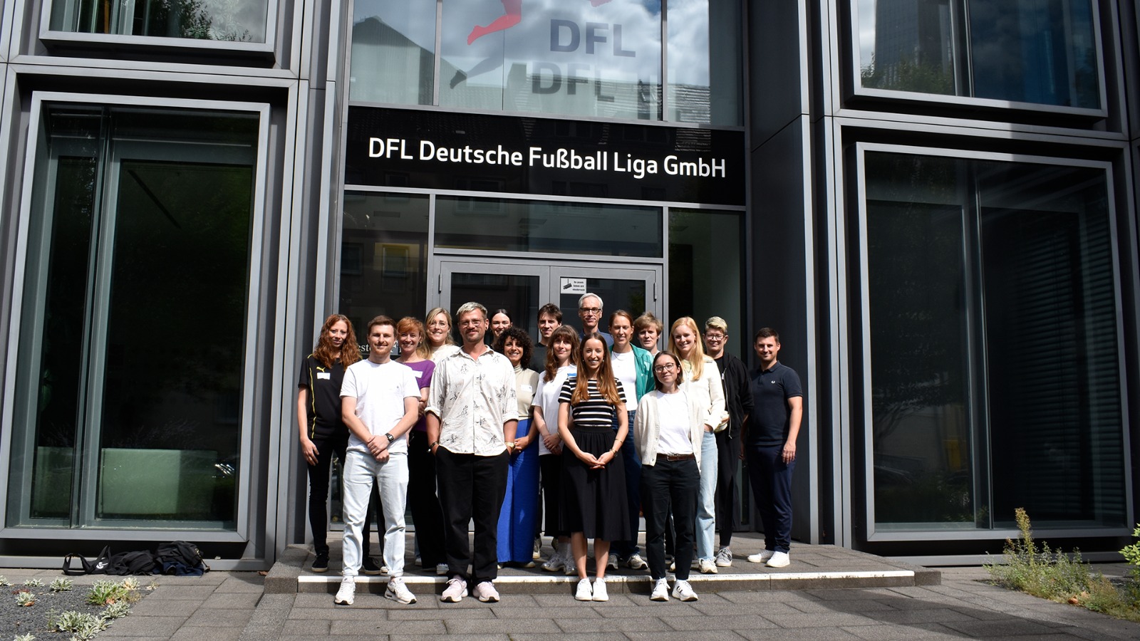 Eine Gruppe von 17 Menschen steht vor der DFL-Zentrale in Frankfurt. Über der Gruppe von Menschen stehen die Worte: “DFL Deutsche Fußball Liga GmbH” mit dem DFL-Logo.
