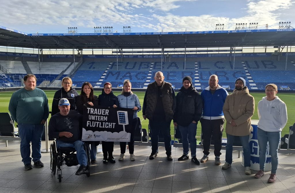 Gruppe von 11 Personen steht im Stadion. Im Hintergrund 1. FC Magdeburg Schriftzug. 3 Personen halten Trauer unterm Flutlicht Banner hoch.