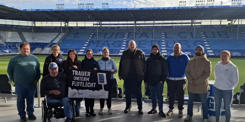 Gruppe von 11 Personen steht im Stadion. Im Hintergrund 1. FC Magdeburg Schriftzug. 3 Personen halten Trauer unterm Flutlicht Banner hoch.