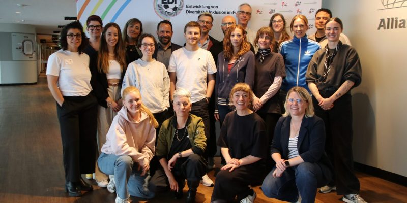 Auf dem Bild sind 20 Personen vor einem EDI Banner zu sehen. Sie lächeln in die Kamera. Die meisten Personen stehen. Davor sind vier Personen in der Hocke. Der Banner ist wie folgt aufgebaut: Links oben sind sechs diagonale Streifen in Regenbogenfarben, rechts daneben das EDI Logo. Rechts daneben die Logos der DFL und von KickIn!