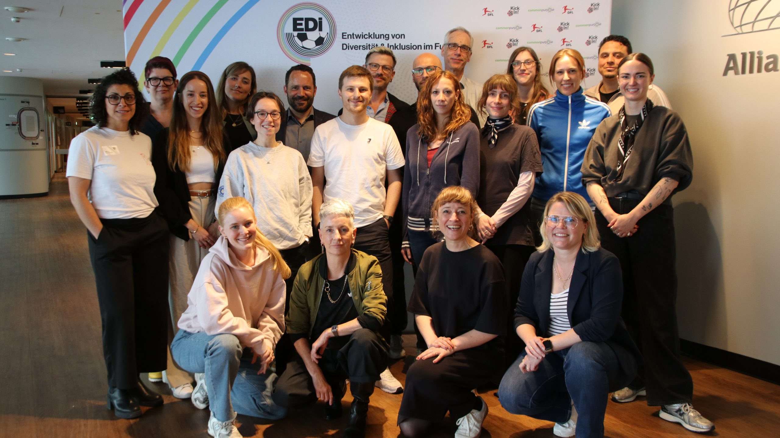 Auf dem Bild sind 20 Personen vor einem EDI Banner zu sehen. Sie lächeln in die Kamera. Die meisten Personen stehen. Davor sind vier Personen in der Hocke. Der Banner ist wie folgt aufgebaut: Links oben sind sechs diagonale Streifen in Regenbogenfarben, rechts daneben das EDI Logo. Rechts daneben die Logos der DFL und von KickIn!